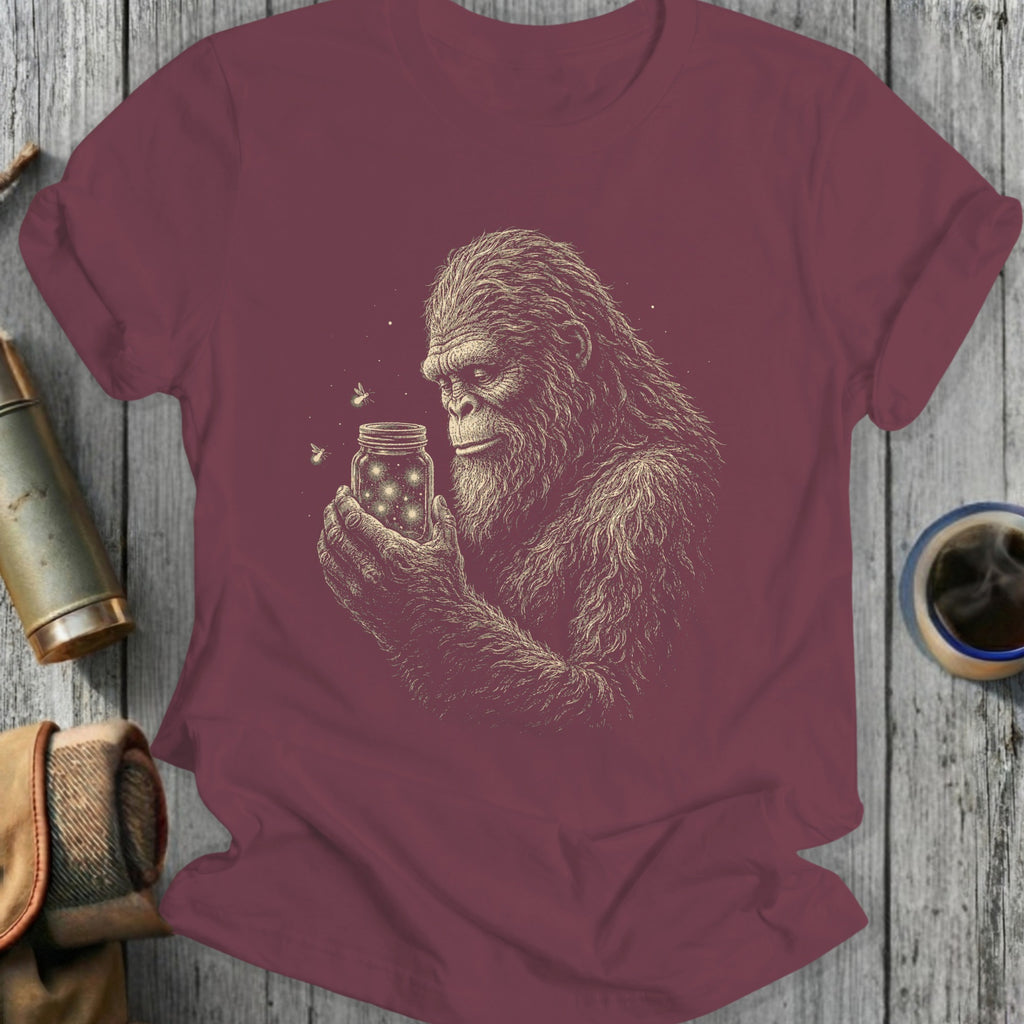 Bigfoot holding a jar of fireflies on a maroon t-shirt.