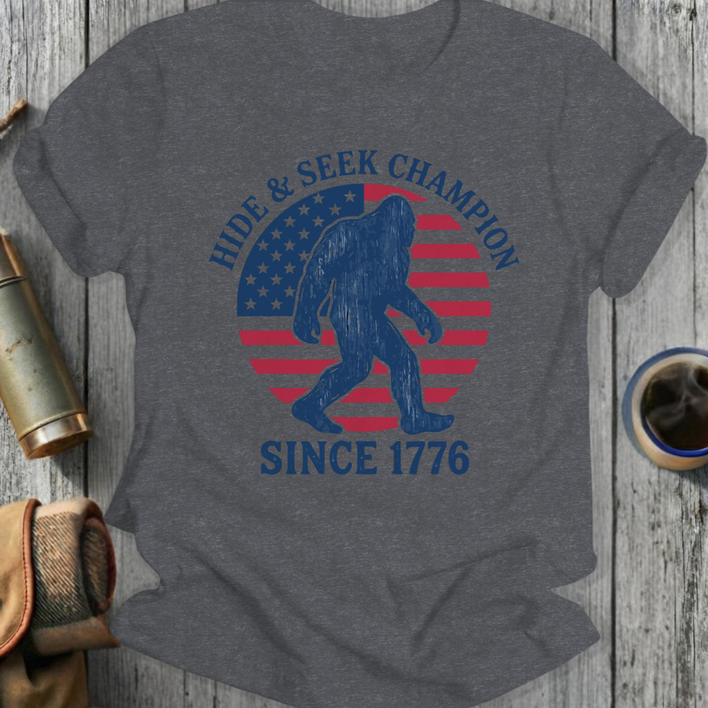 Graphic t-shirt with Bigfoot silhouette, American flag, and text "Hide & Seek Champion Since 1776".