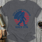 Graphic t-shirt with Bigfoot silhouette, American flag, and text "Hide & Seek Champion Since 1776".