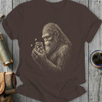 Bigfoot holding a jar with fireflies, drawn in sketch style on a brown t-shirt.