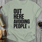Light green t-shirt with bold black text "OUT HERE AVOIDING PEOPLE" and a small pine tree icon.