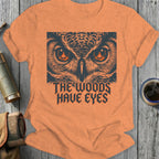 Orange t-shirt with graphic of owl eyes and text "The Woods Have Eyes".
