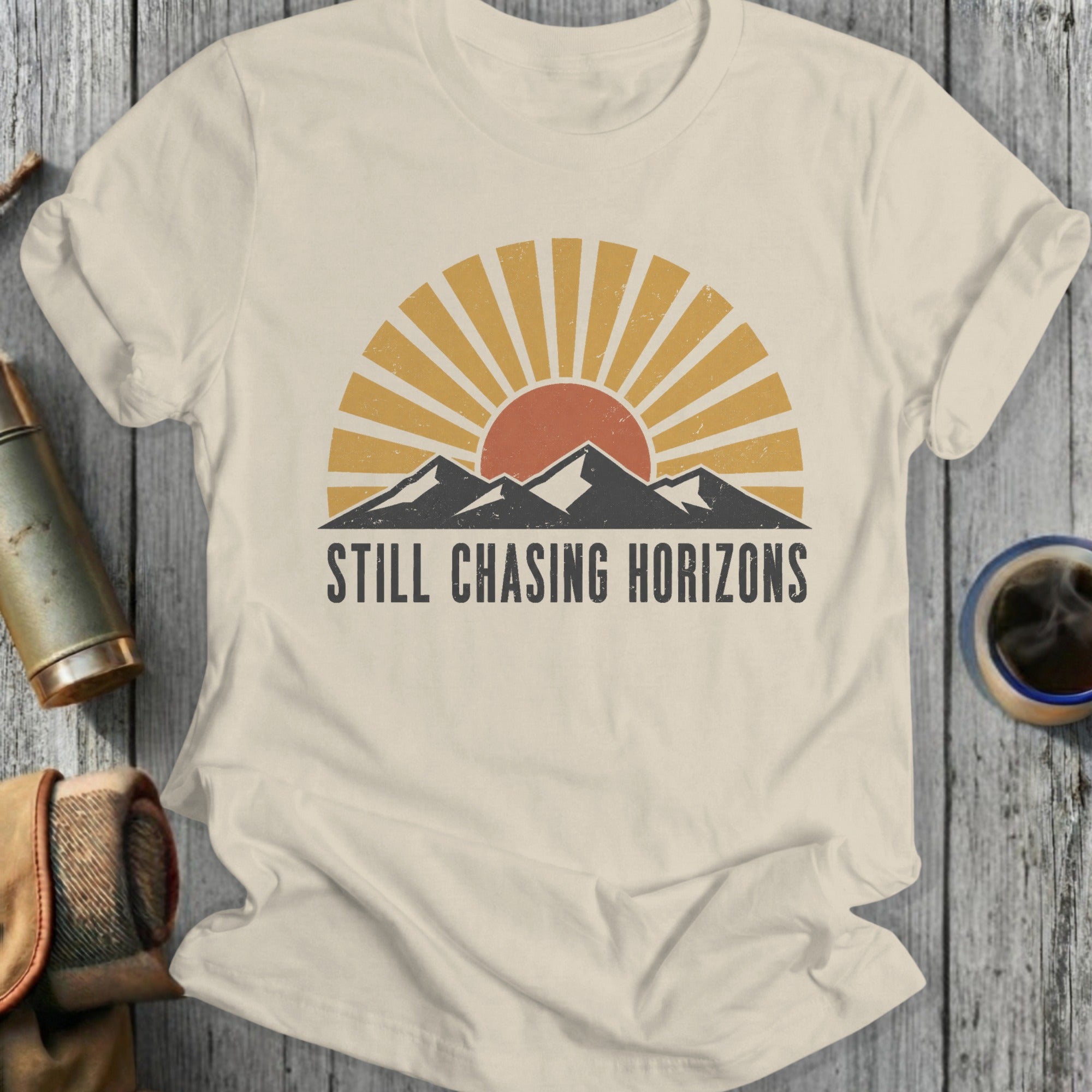 Cream t-shirt with sun, mountains, and "STILL CHASING HORIZONS" text.