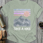 T-shirt with watercolor mountains, pink sun, striped sky, and "TAKE A HIKE" text.