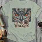 T-shirt graphic with owl eyes, campfire flames, and text "The Woods Have Eyes."