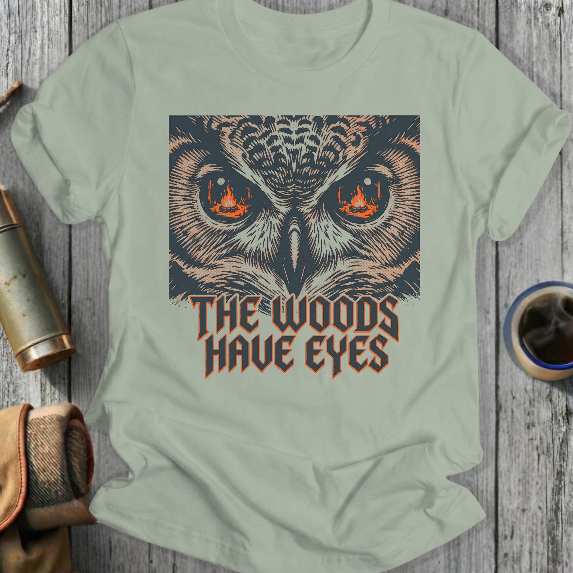 T-shirt graphic with owl eyes, campfire flames, and text "The Woods Have Eyes."