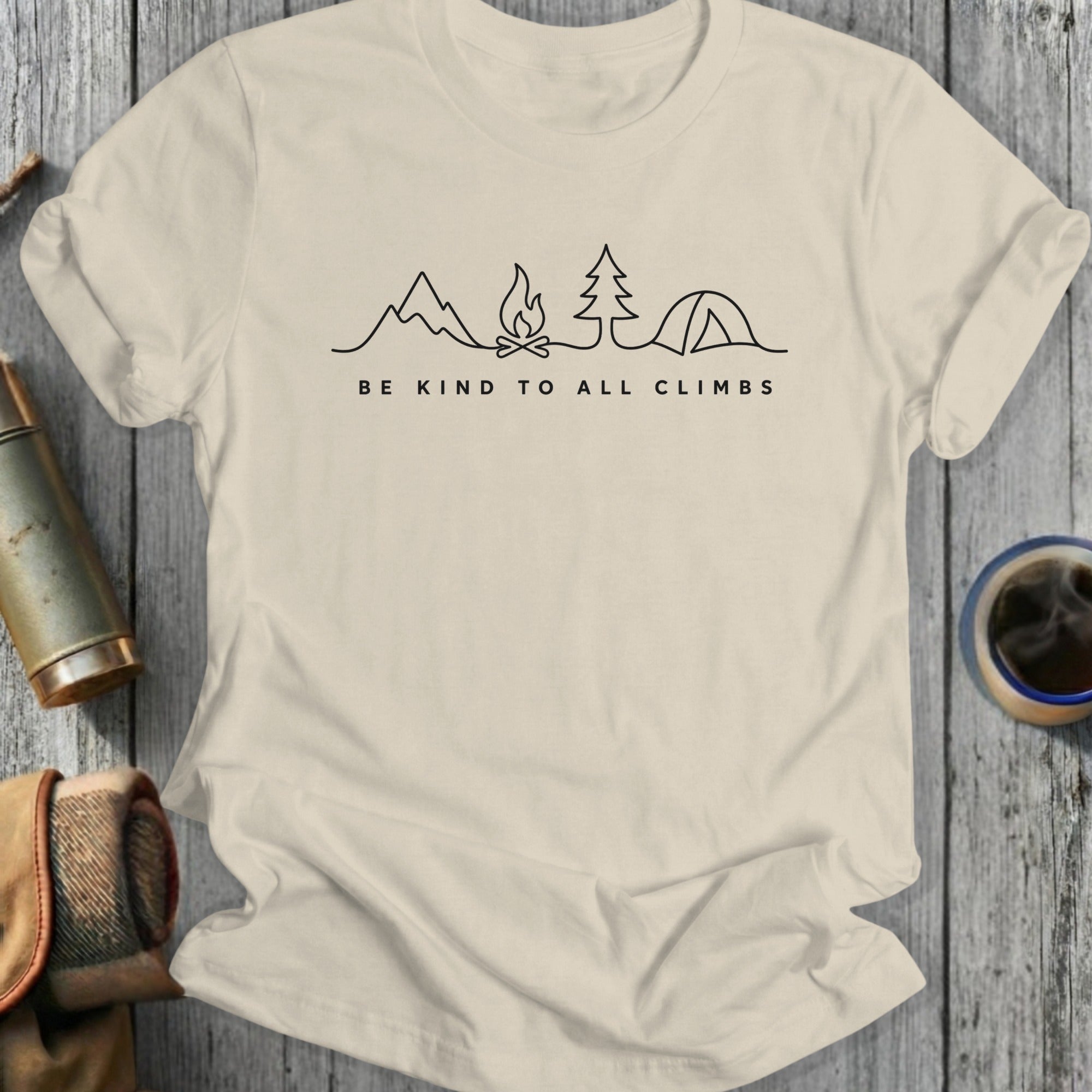 T-shirt with minimalist mountain, campfire, tree, and tent illustration and "Be Kind to All Climbs" text.