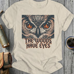 T-shirt graphic featuring an owl's face with fiery eyes and text reading "The Woods Have Eyes."