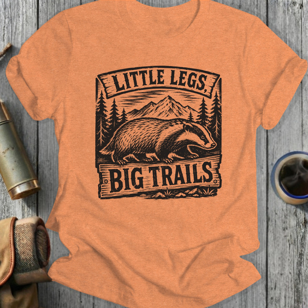 T-shirt graphic with badger, mountains, trees, and text "LITTLE LEGS BIG TRAILS".