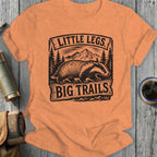 T-shirt graphic with badger, mountains, trees, and text "LITTLE LEGS BIG TRAILS".
