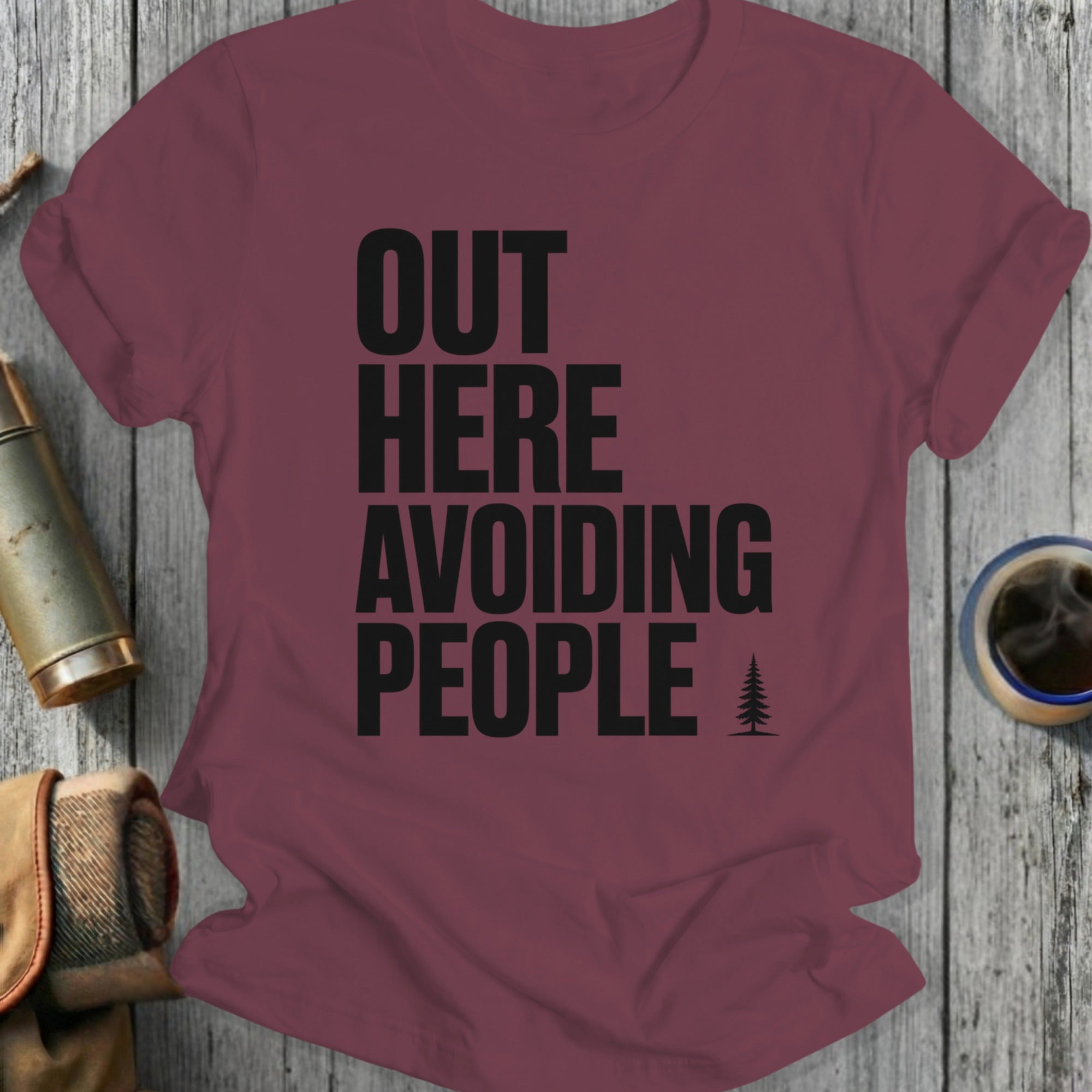 Burgundy t-shirt with black text "OUT HERE AVOIDING PEOPLE" and a small tree icon.