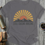 Grey t-shirt with sun, mountains, and "STILL CHASING HORIZONS" text.