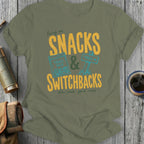Olive t-shirt with yellow text "Snacks & Switchbacks" and trail mix graphics.