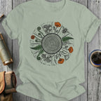 Light sage t-shirt with a circular woodland illustration of a tree ring, ferns, mushrooms, and a bee.
