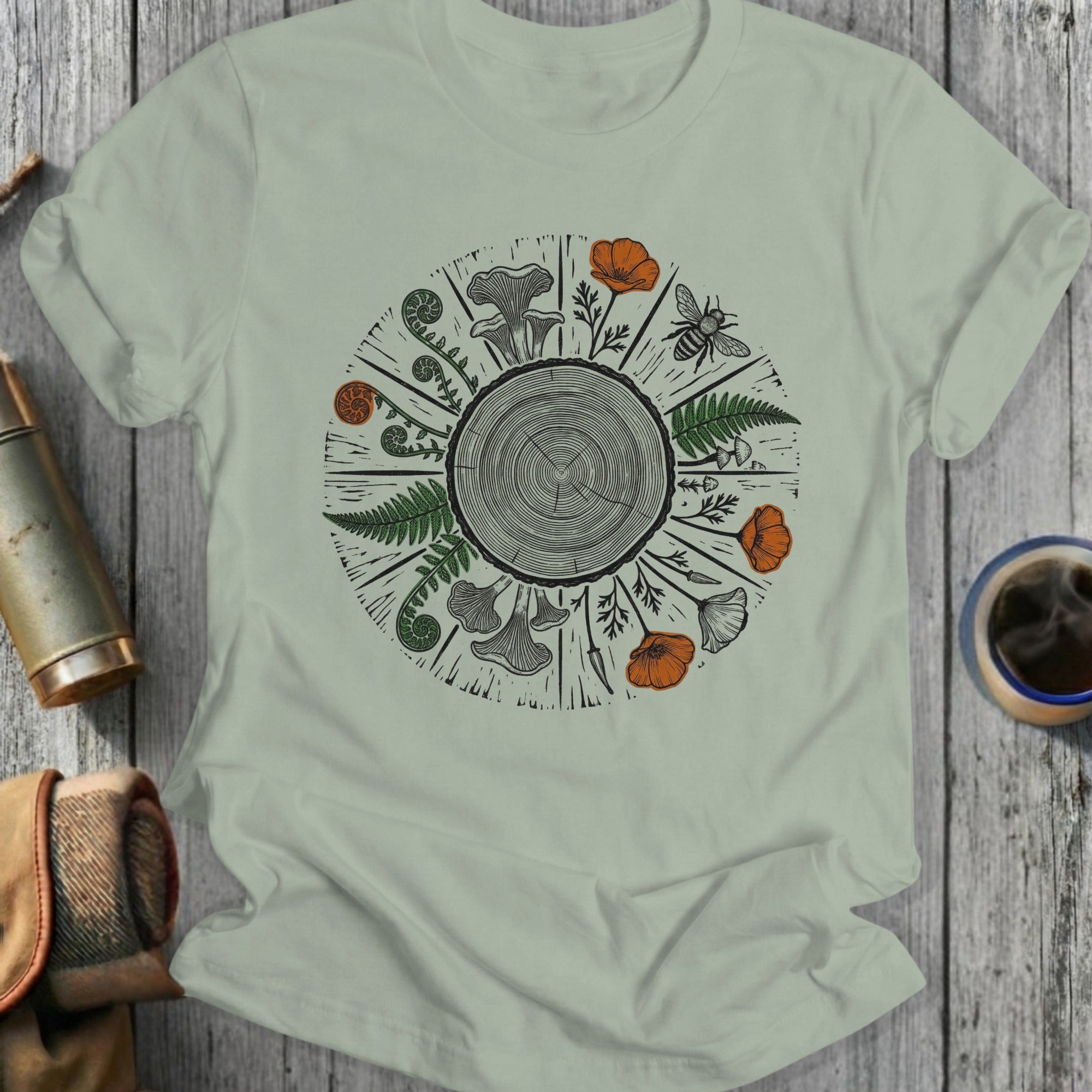 Light sage t-shirt with a circular woodland illustration of a tree ring, ferns, mushrooms, and a bee.