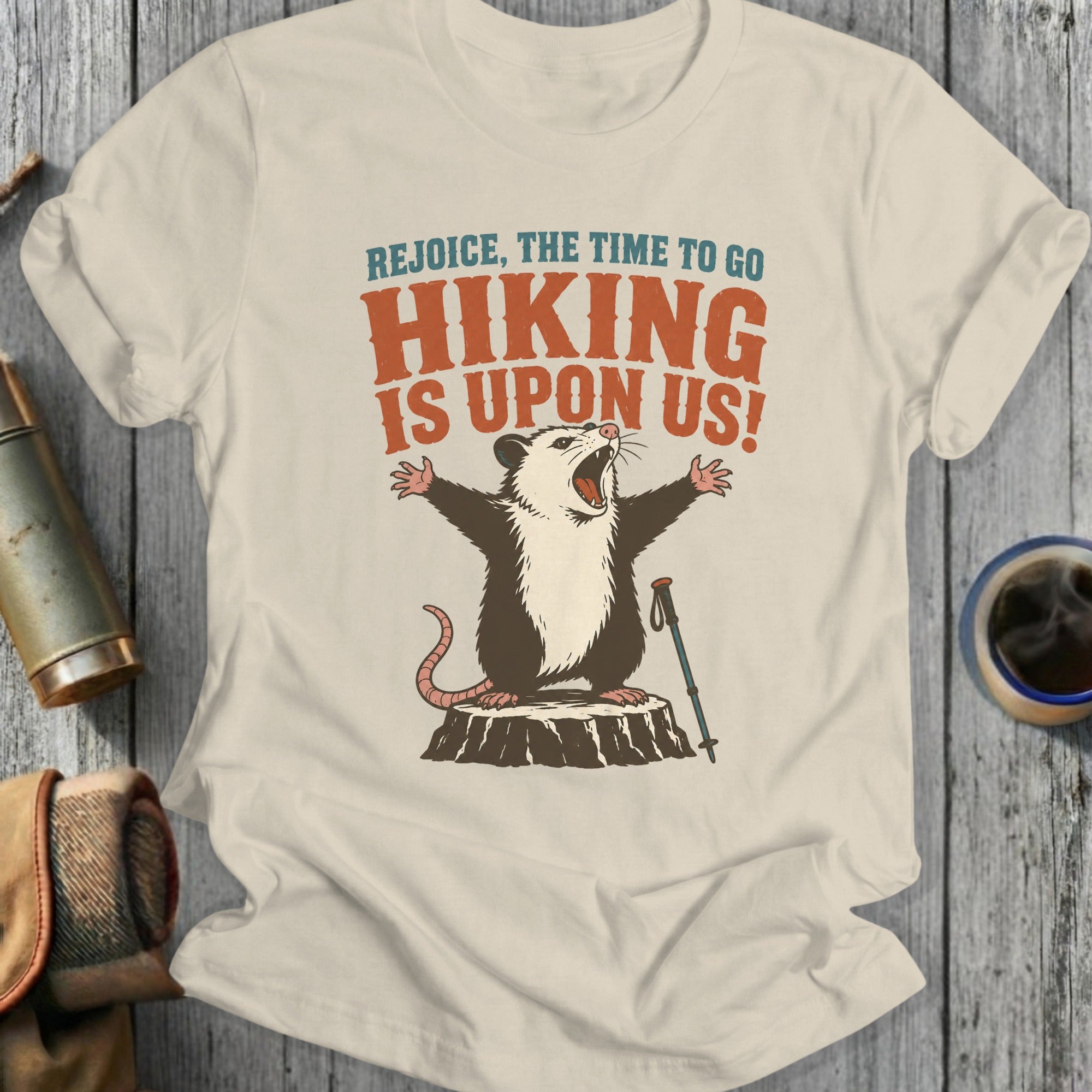 Opossum with arms outstretched on a tree stump with hiking pole, "Rejoice, The Time To Go Hiking Is Upon Us!" text.