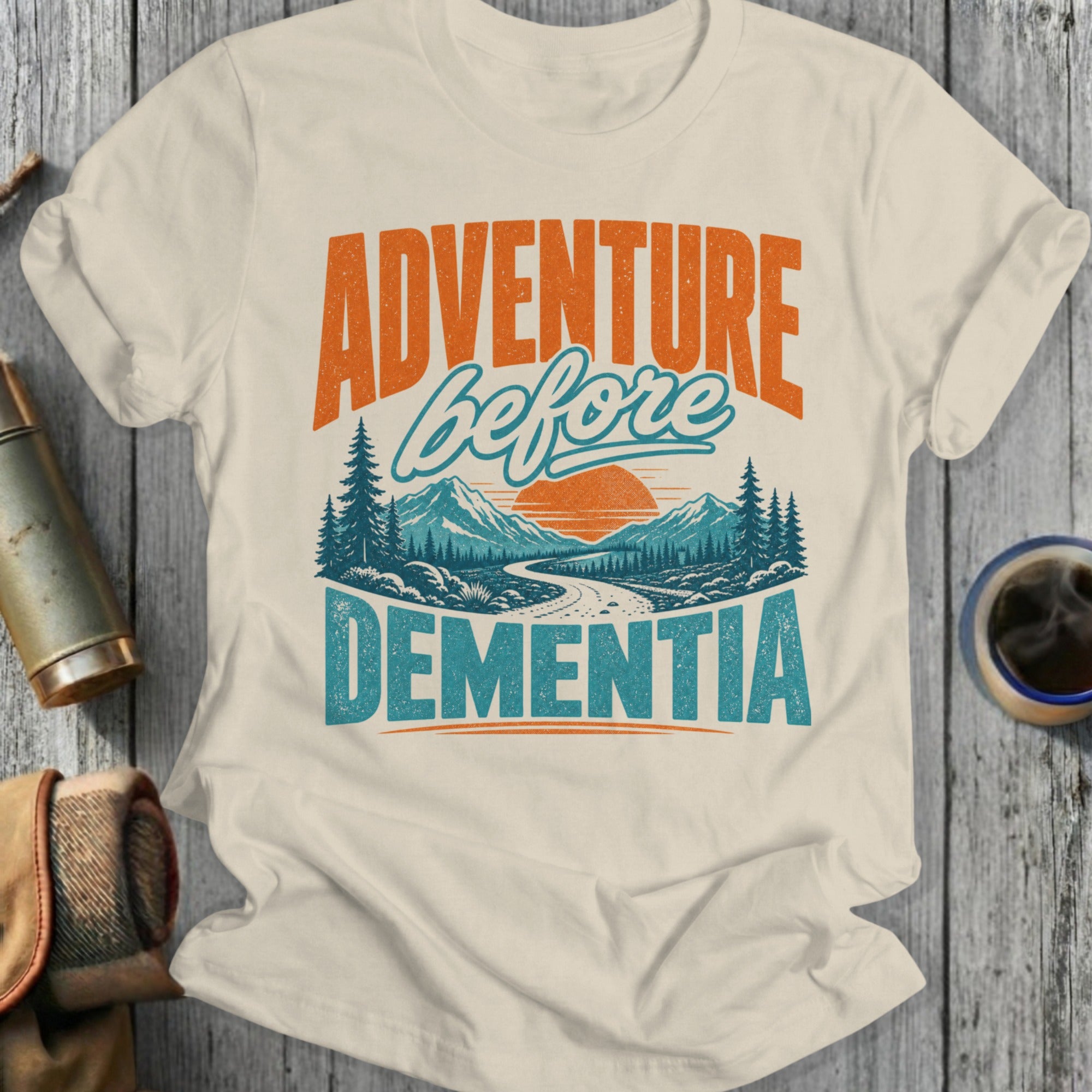 Cream t-shirt with "ADVENTURE before DEMENTIA" text and mountain landscape design.