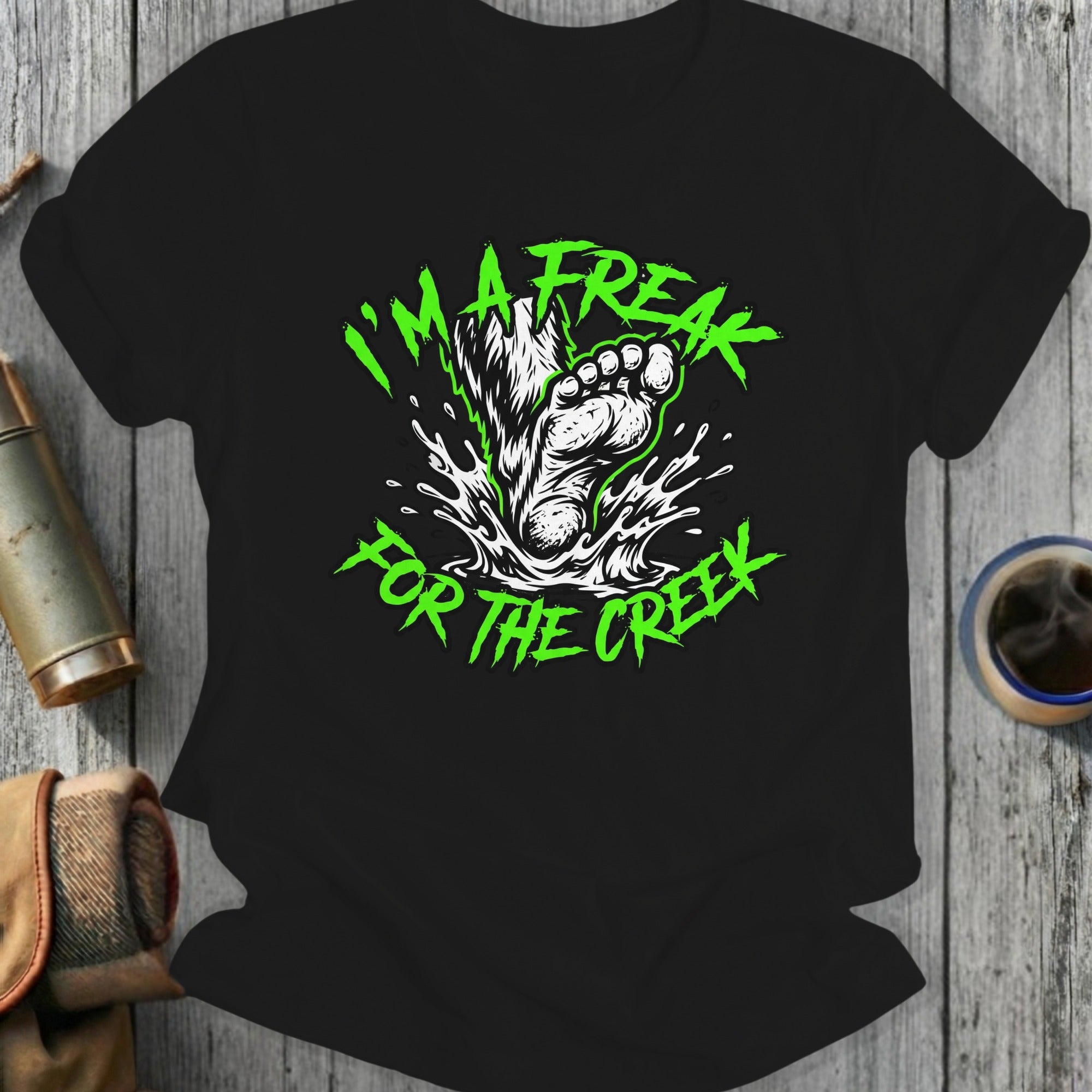 Black t-shirt with green "I'm a freak for the creek" graphic featuring Bigfoot's foot splashing water.