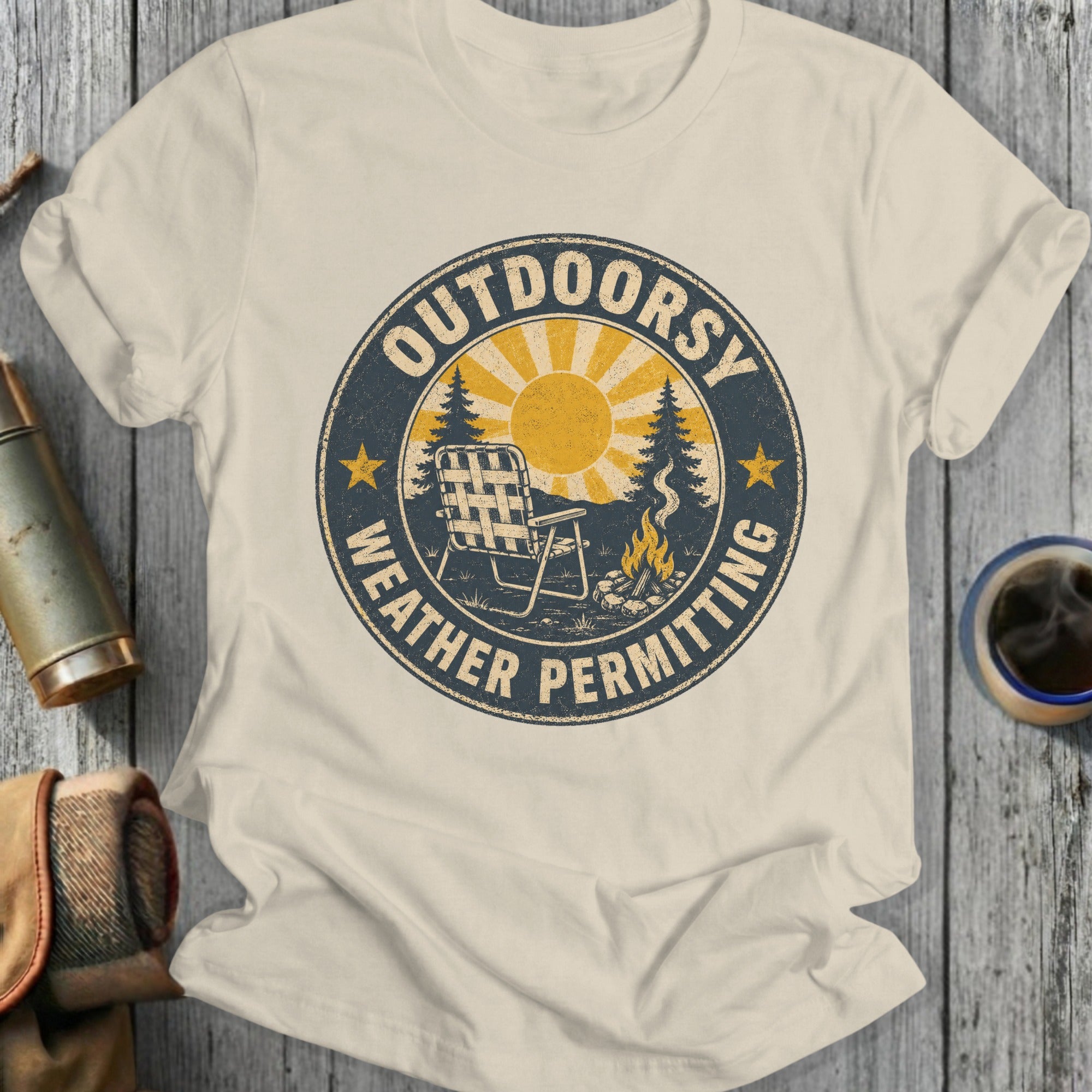 Cream t-shirt with distressed circular graphic of chair, campfire, and sun with text "OUTDOORSY WEATHER PERMITTING".