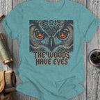 T-shirt graphic featuring owl eyes with fire and text "THE WOODS HAVE EYES".