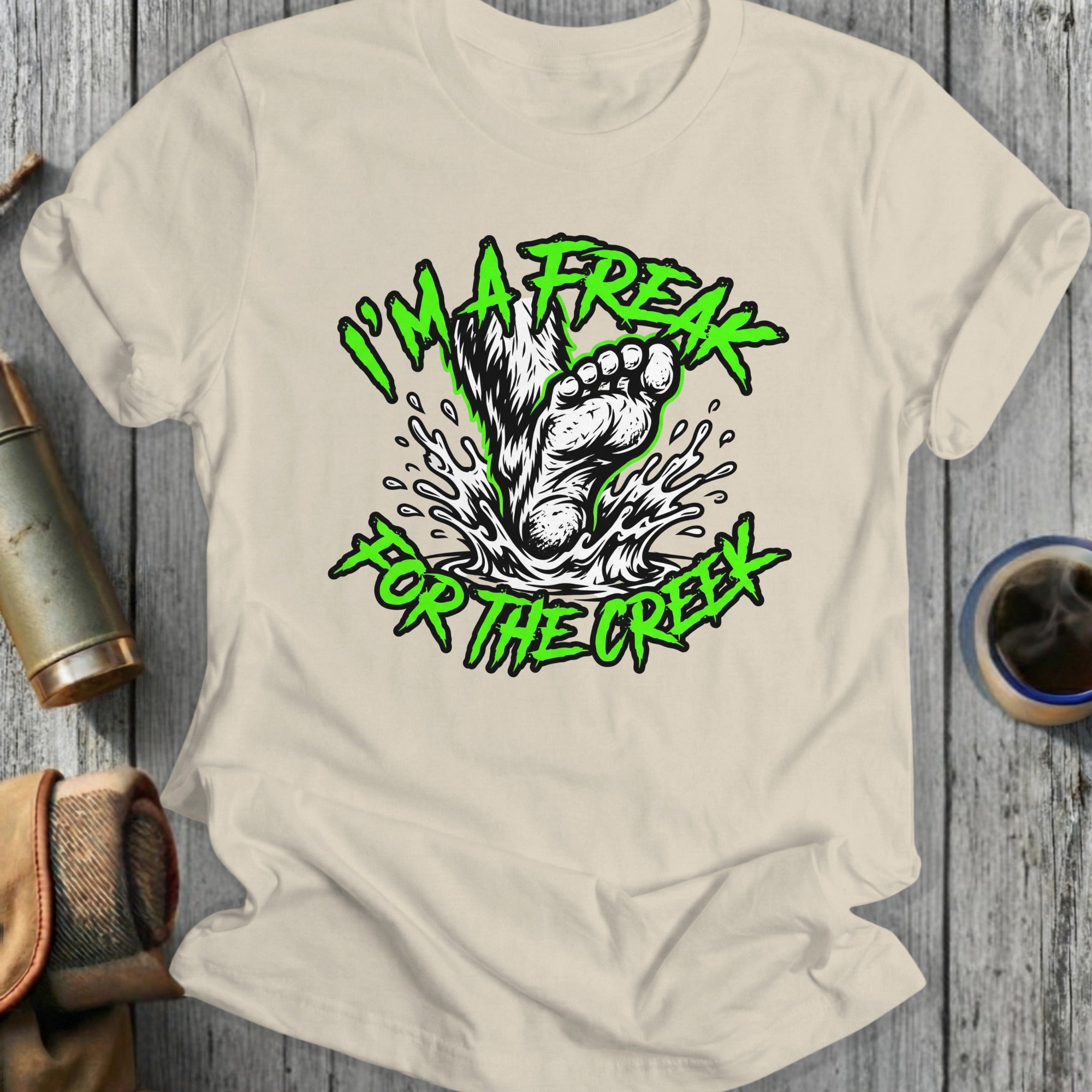 Green text and Bigfoot foot splashing water on cream t-shirt.