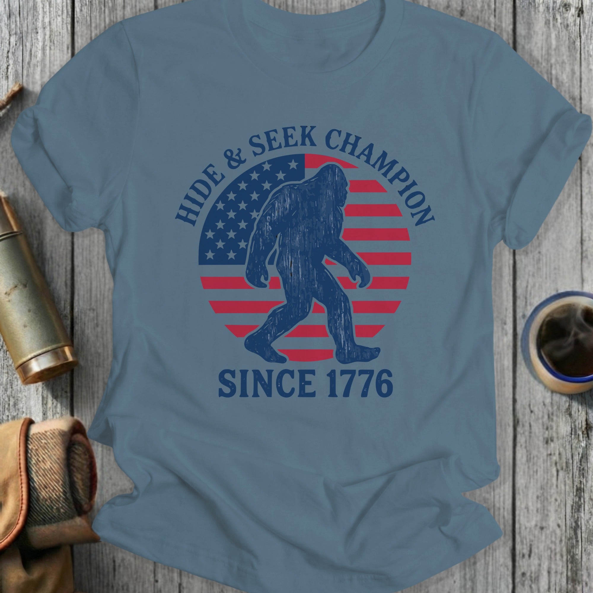 Blue t-shirt with Bigfoot silhouette, American flag, and text "HIDE & SEEK CHAMPION SINCE 1776".
