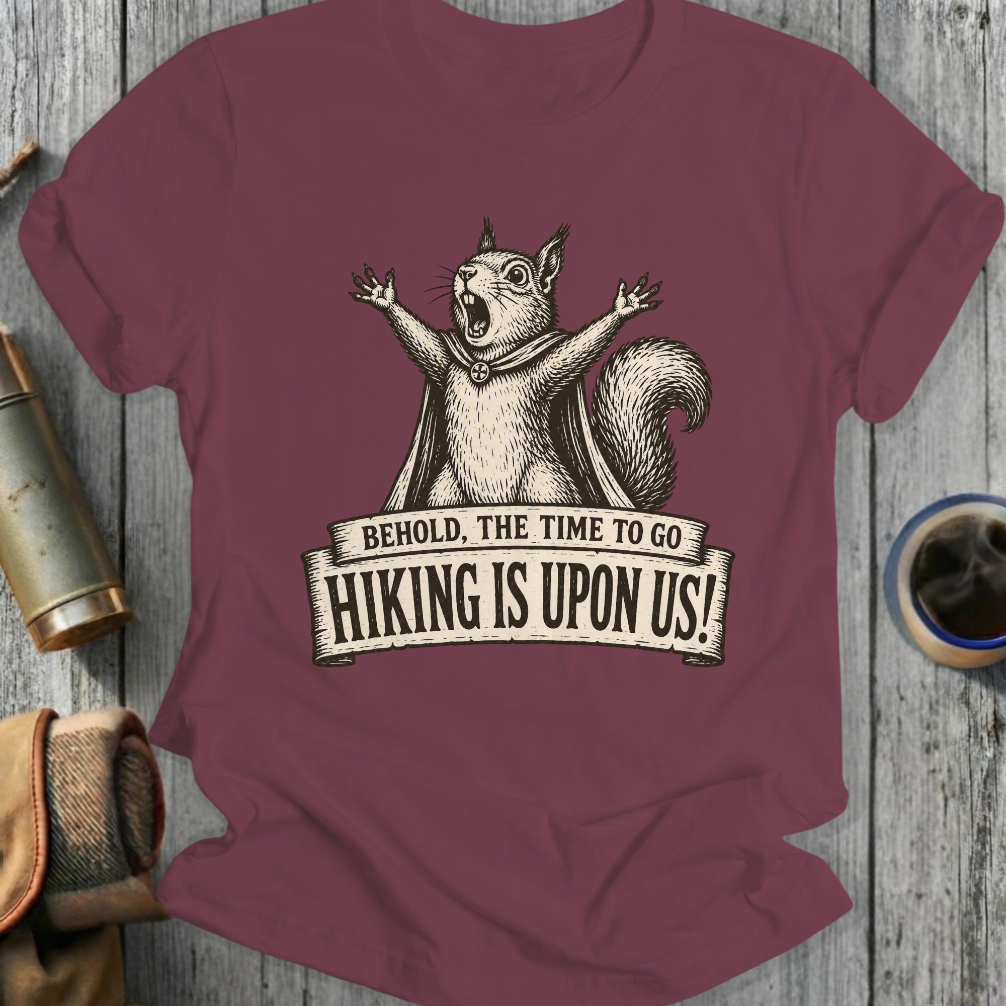 Maroon t-shirt with a distressed print of a squirrel wearing a cape and the text "BEHOLD, THE TIME TO GO HIKING IS UPON US!".