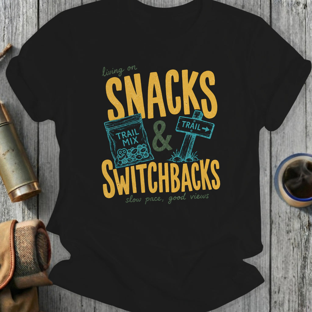 Black t-shirt with "Snacks & Switchbacks" graphic, trail mix bag, and trail sign.