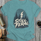 Teal t-shirt with "STILL FERAL" text, forest graphic, and lightning bolt.