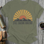 Olive green t-shirt with "STILL CHASING HORIZONS" text and sunset mountain graphic.