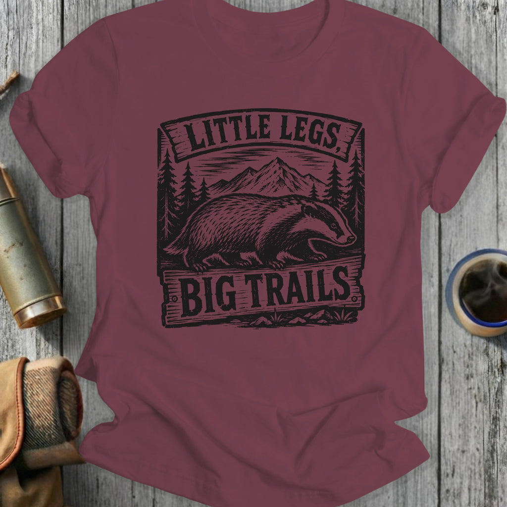 Maroon t-shirt with bear graphic and 'Little Legs Big Trails' text on a wooden surface.