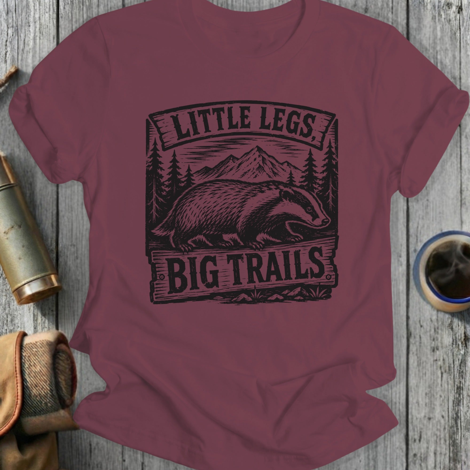 Maroon t-shirt with bear graphic and 'Little Legs Big Trails' text on a wooden surface.